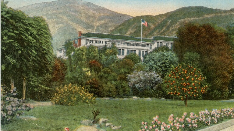 A hand-colored postcard of a large, white, colonial-style building with a green tiled roof stands behind a lush landscape of flower beds, a green lawn and many varieties of trees, with mountains looming just behind. An American flag waves at the top of a flagpole above the roof.