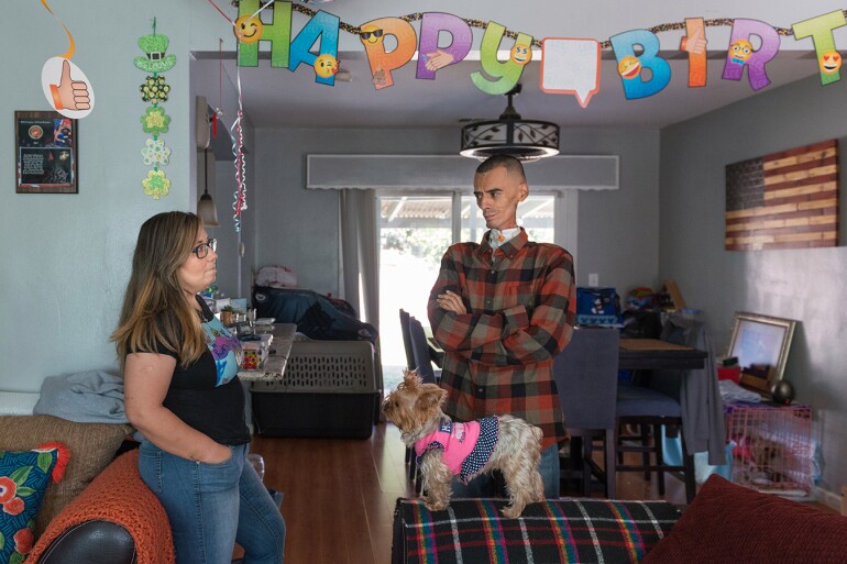 Rocio and Brian stand in the living room of their home. The walls are painted in a powder blue color. To the right, there is an American flag wall art. Above them hangs a "Happy Birthday" banner.
