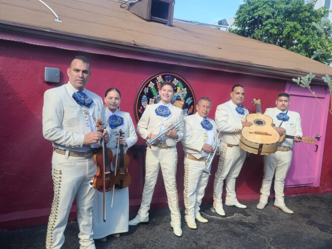 Mariachis con trajes blancos portando sus instrumentos frente a una casa roja