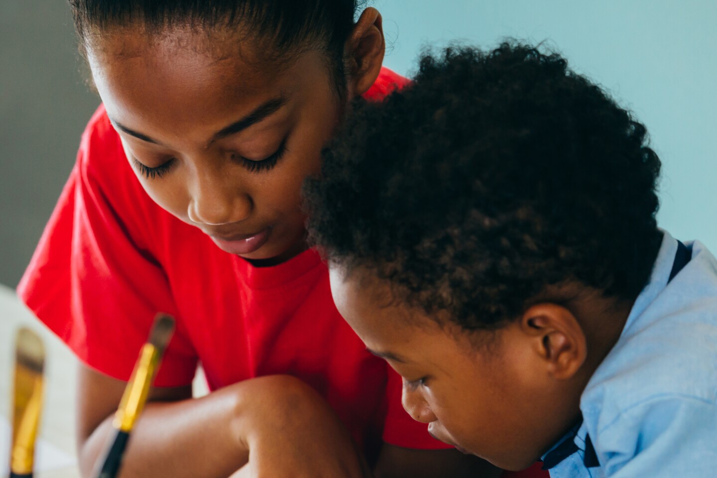 Two Black children lean into something they are working on on a table with paintbrushes and crayons.