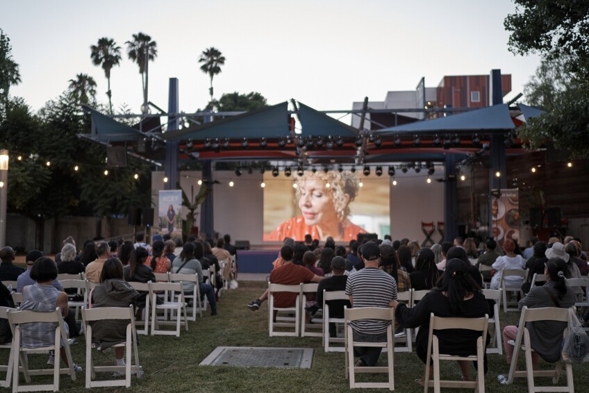An outdoor audience sits in white outdoor chairs as everyone faces a screen projecting an episode of "The Great American Recipe."