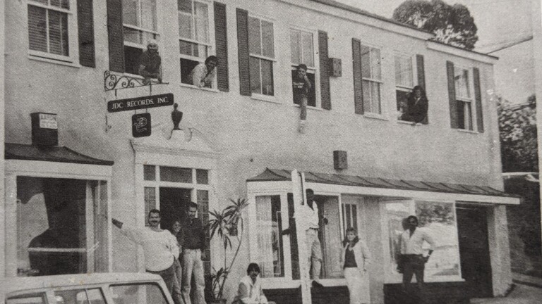 A black and white photo of a two-story building with a sign hanging off the side that reads, "JDC Records Inc." Men and women are posed in front of the building and poking their heads out of the windows and doors.