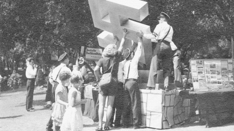 An archival black and white photo depicts people raising a large Swastika sculpture on a pedestal at a park.