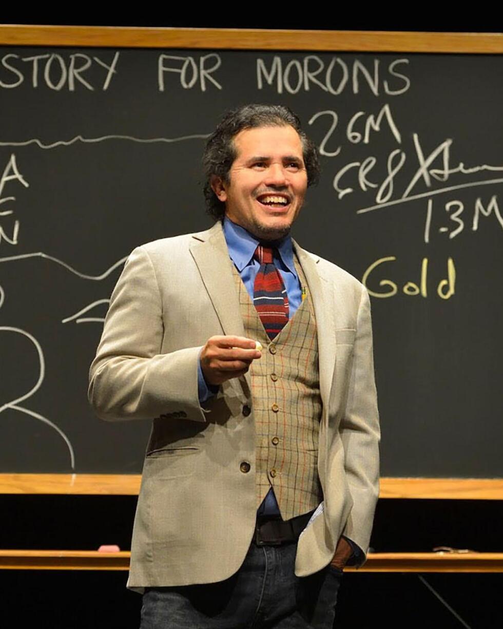 John Leguizamo on stage stands in front of a chalkboard that reads "Latin History for Morons."