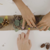 A child's hands and an adult's hands attach natural items to a paper headband.