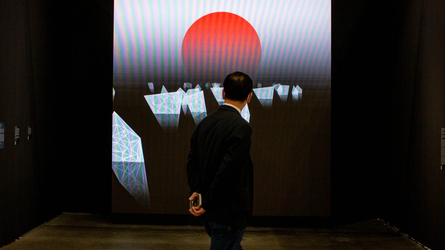 A man in a suit with his hands behind his back looks on to a digital art piece on a large LED screen mounted on a black gallery wall. The digital art piece features a large red dot resembling a setting sun with floating white "icebergs" on a black water surface.