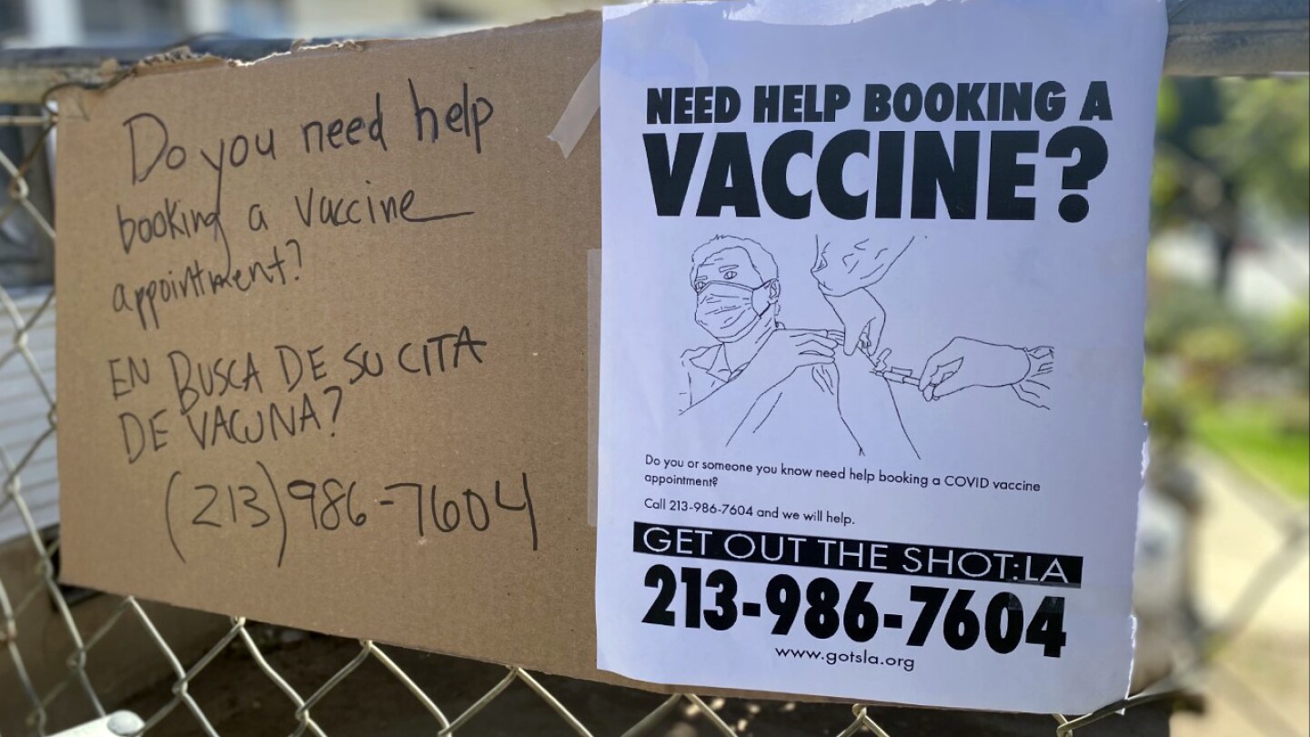 A cardboard and paper sign has a picture of a person being vaccinated and reads "Need help booking a vaccine appointment?" and lists the number 213-986-7604 and the website www.gotsla.org.