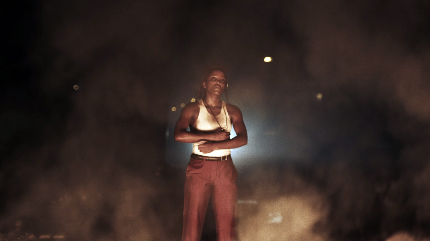 Still from Yere Faga: A woman stands behind flames and smoke at night.