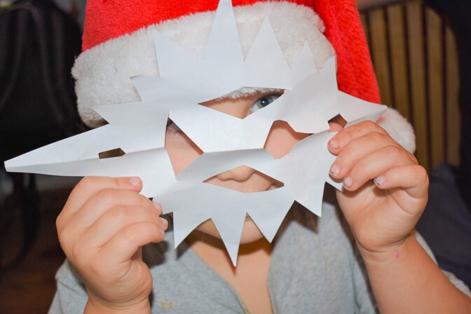 A small child wearing a red Santa hat holds a paper snowflake in front of the face like a mask.