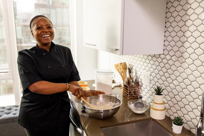 Opportune Akendeu is wearing a completely black chef's suit. She is standing in a kitchen next to a counter where she mixes a metal bowl with puff dough. She stirs the dough with a wooden spoon and smiles at the camera.