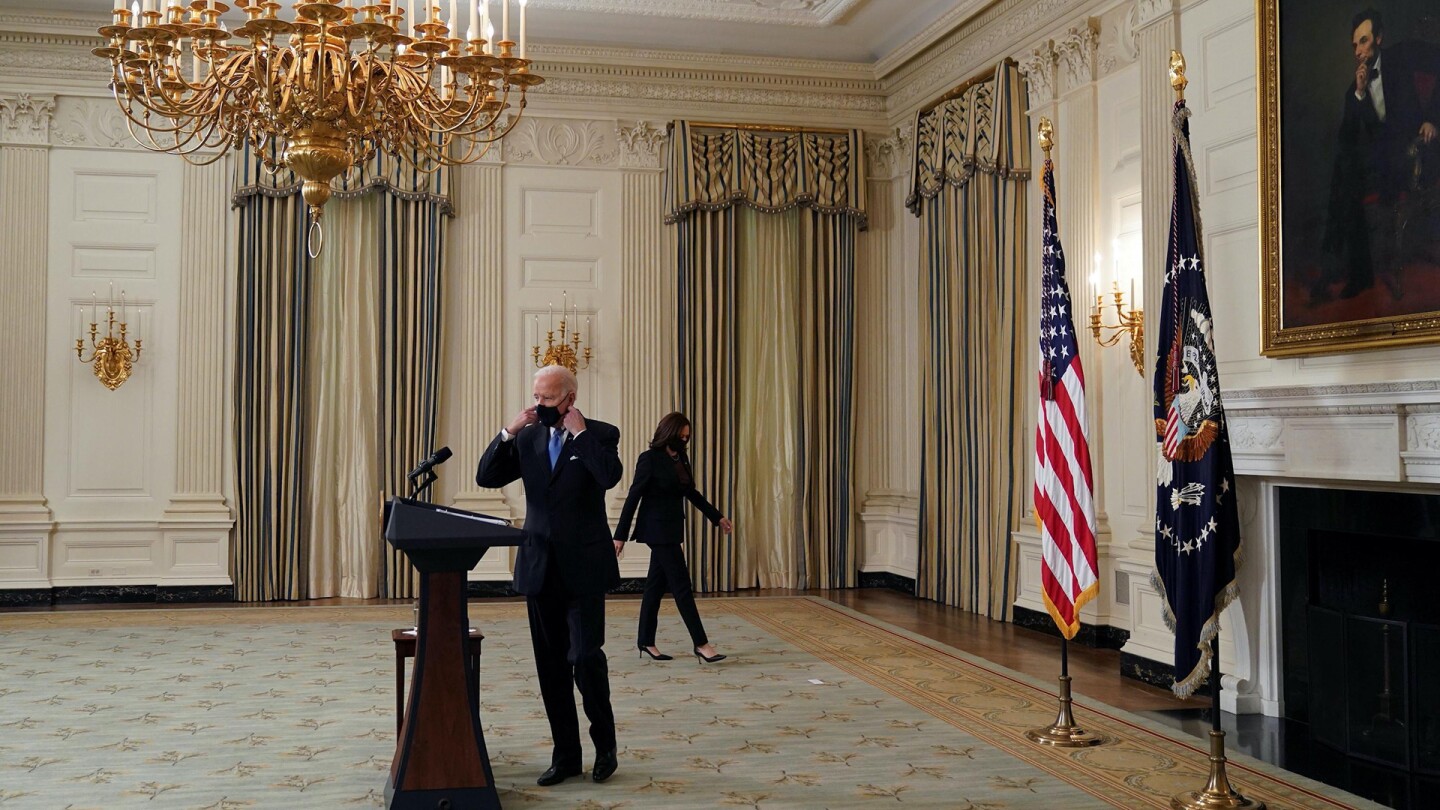 President Joe Biden taking off his face mask on his way to a lectern, with Vice President Harris behind him.