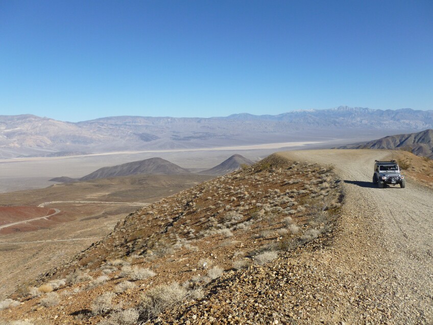 A silver jeep sits on a dirt path with a wide view of Death Valley behind it.