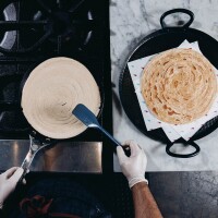 An overhead shot of lachha paratha dough is heated on a skillet over a stove. To the right of the stove are a stack of completed lachha parathas. 