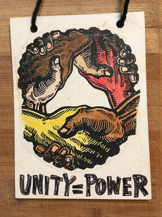 A mat board necklace with a circle of multicolored hands holding each other that reads "Unity=Power"