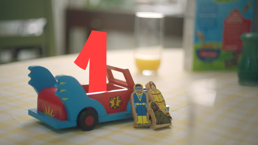 A small toy car and 2 toy figures are arranged on a table. A large red 1 hovers inside the car.