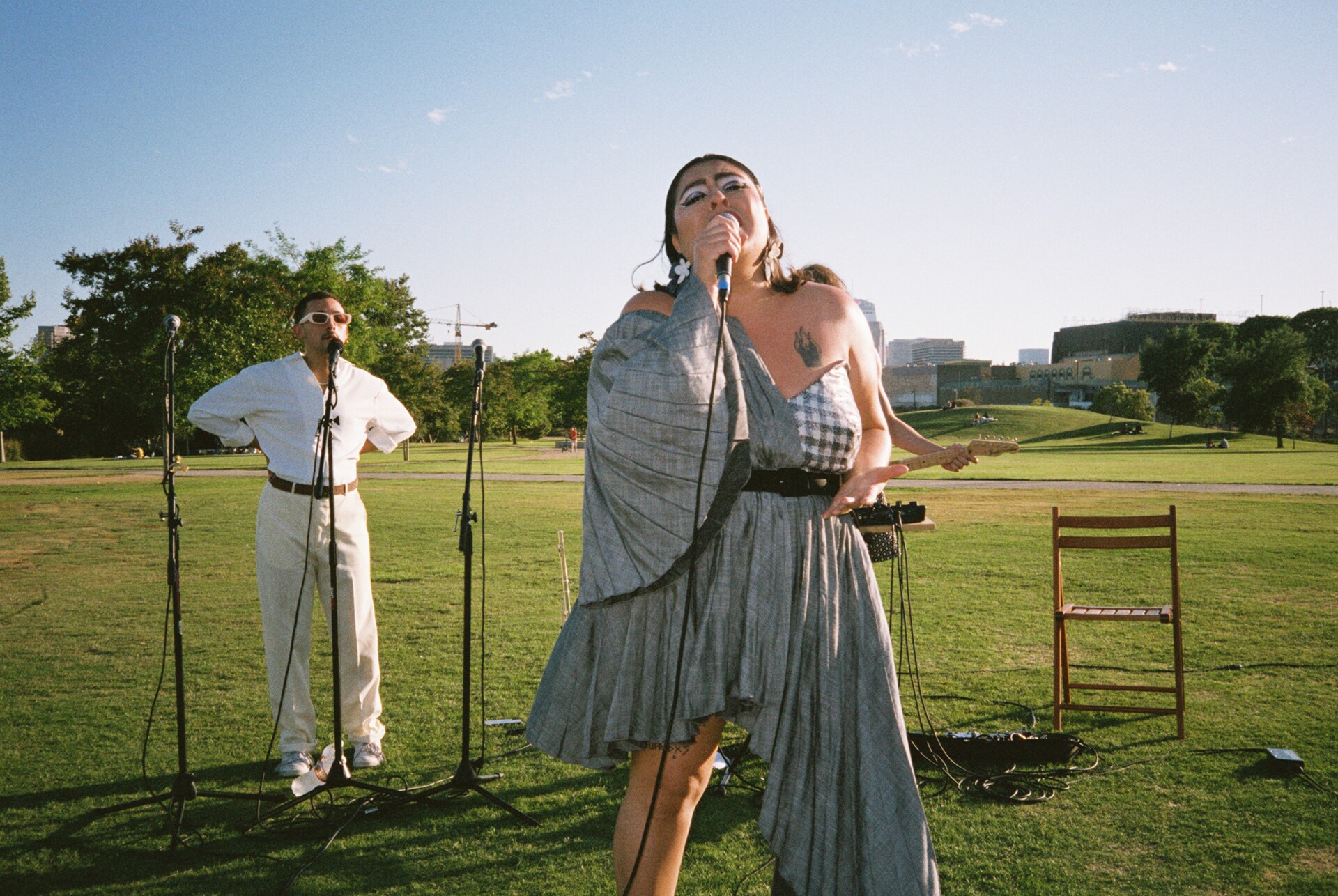 San Cha is standing and singing into a microphone. They are wearing big, white statement earrings, an asymmetrical silver dress that flares in the arms and exposes her shoulder, one of which has a tattoo. Behind them is another band member wearing white and singing into a microphone stand with their hands on their hips. They are wearing white sunglasses. The entire band is playing on an open grassy field.