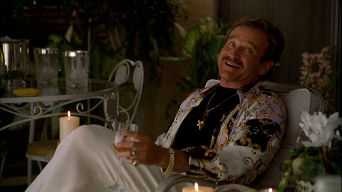 Robin Williams leans back on a chair holding a drink glass.