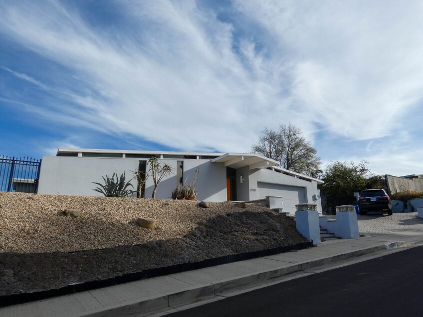 A white house with a slanted roof. The slant is a low slope. The front door is a brown-red color. The house sits on a hill and has a driveway where an SUV is parked.  