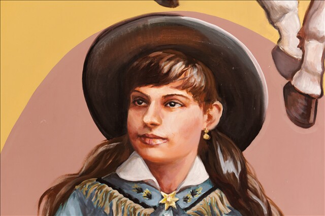 A painting of Annie Oakley in a close-up of the "Spirits of the West" mural. Oakley is wearing a wide-brimmed hat and has long, dark brown hair that flows behind her. She's wearing a collared blue top with a white collar and beige fringe. A gold star broach is pinned in the middle.
