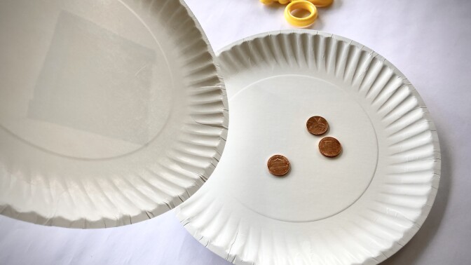 One empty paper plate is tilted over another that holds three pennies on a table.