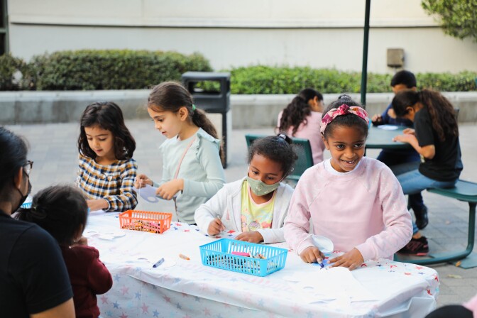 Children gather around outdoor tables and use crayons to color white paper. Colorful plastic bins full of crayons are placed at the middle of the table.