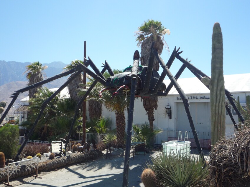 A large spider sculpture, with the body of the spider replaced with a full-sized Volkswagen Beetle, towers over a walkway leading up to a building with a sign that reads, "Hole in the Wall" over the door. 