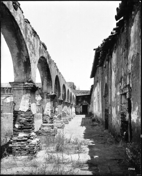 Photograph of a view of the corridor at Mission San Juan Capistrano, as seen from the west side of the mission, Orange County, California, 1885. Wild, tall grass grow cover the open space (quadrangle?) to the left. Several patches of the same grass are present in the area in the foreground. The corridor of what once was an arcade is in dilapidated condition. Parts of the stucco are worn off revealing the deteriorating adobe bricks. The buildings in the background and to the right are in similar conditions.