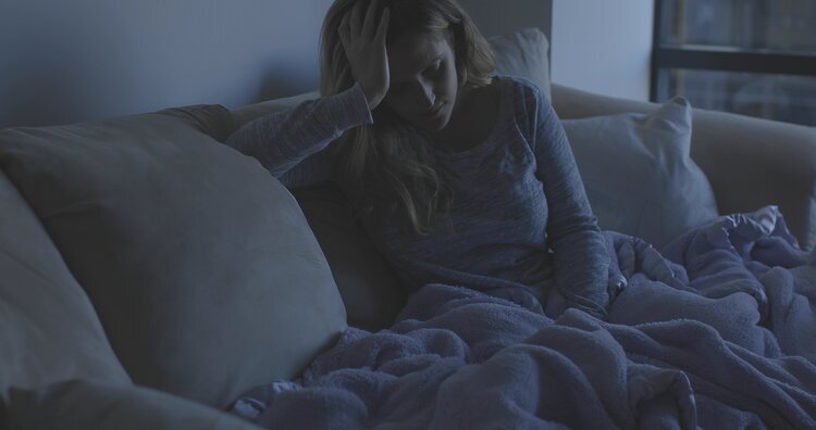 Jennifer sits on her couch in a dimly lit room. She's resting her head on her hand, propped up by the couch. A blanket covers her legs and feet.