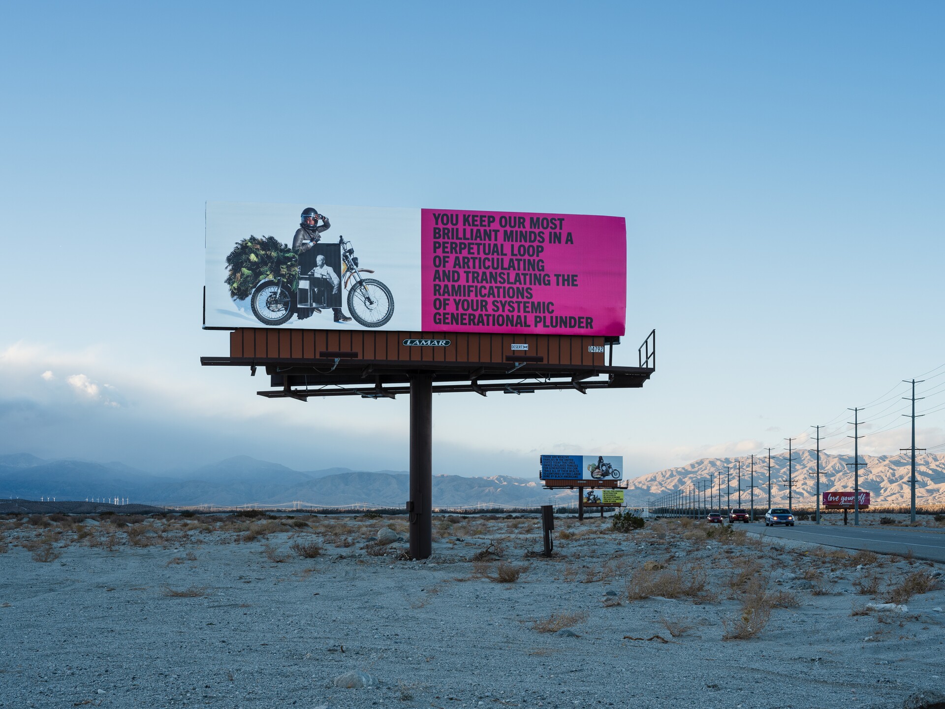 Simmons' billboards stand along the Gene Autry Trail. The one photographed reads, "You keep our most brilliant minds in a perpetual loop of articulating and translating the ramifications of your systemic generational plunder."