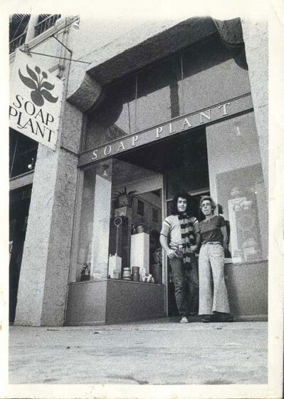 An archival black and white photo of Billy Shire and his mother, Barbara Shire, standing in front of their family-store, Soap Plant. The store has a glass storefront with the words "SOAP PLANT" written across and a store sign reading the same thing.
