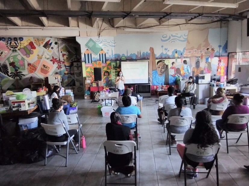 Plastic chairs are arranged in a room spaced apart. People sit in them and face the front where a woman is standing in front of a projector, speaking to the group. Behind the presenter is a colorful mural.  