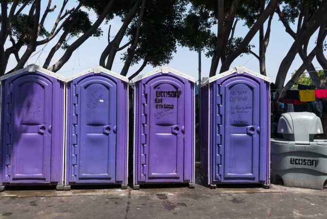 Four purple portable toilets stand side by side. Some have various writing scrawled on them in black ink.