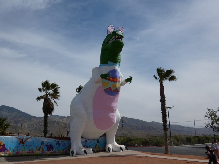 A three-story concrete T. Rex painted from neck down to resemble a bunny. A blue bow tie is painted around its neck and an easter egg painted on its chest. Bunny ears are placed on the dinosaur's head. 