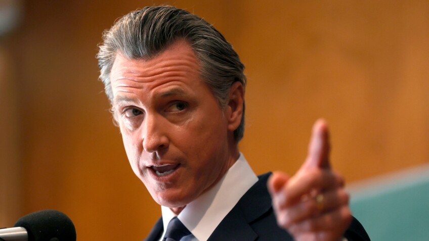 A Caucasian man with slicked-back salt-and-pepper hair stands behind a microphone and looks off to his left as his mouth is open mid-sentence and he points with his left index finger in the same direction.