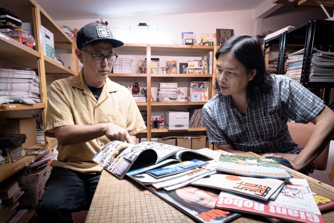 Two men are seated in front of a table filled with magazines that read "Giant Robot" on each cover. One man is holding an open magazine and pointing at something inside while the other man leans over the table to look. They are in what appears to be a closet, surrounded by shelves filled with stacks of magazines and other mementos. 