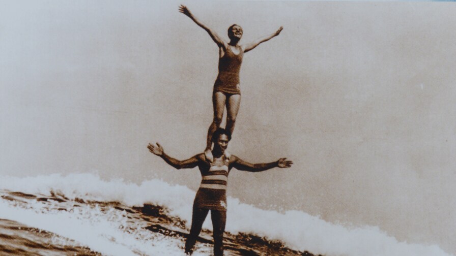 A sepia-toned photo of Duke Kahanamoku surfing a wave while Viola Hartman stands on his shoulders. The two have their arms outstretched as they ride the wave.