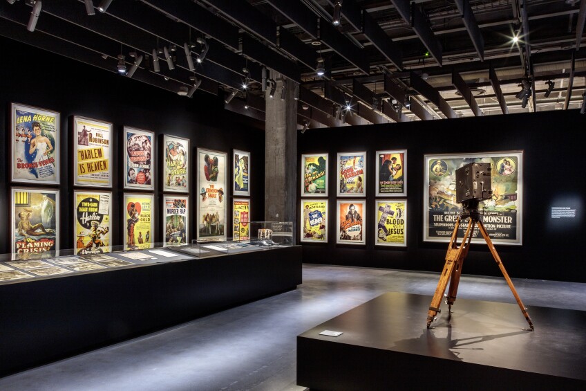 Two museum gallery walls are filled with movie posters of race films, arranged in a grid. In the foreground, a vintage movie camera is propped on a tripod and placed on a pedestal. Photographs and other exhibition materials are arranged in a glass case. 