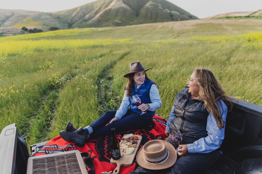 Mireia Taribó, left, and Tara Gomez, right, of Lompoc's Camins 2 Dreams, sit in the bed of a truck parked in a lush green field. They're seated on a tribal print red and black blanket with a charcuterie board in between them. They enjoy a bottle of wine together.