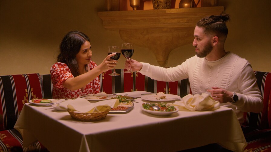 Natasha, who is wearing a red dress with white flowers, and Shant, who is wearing a white, cable-knit sweater, clink wine glasses over a dinner table topped with a white tablecloth and various dishes. Natasha and Shant look at each other as they toast.