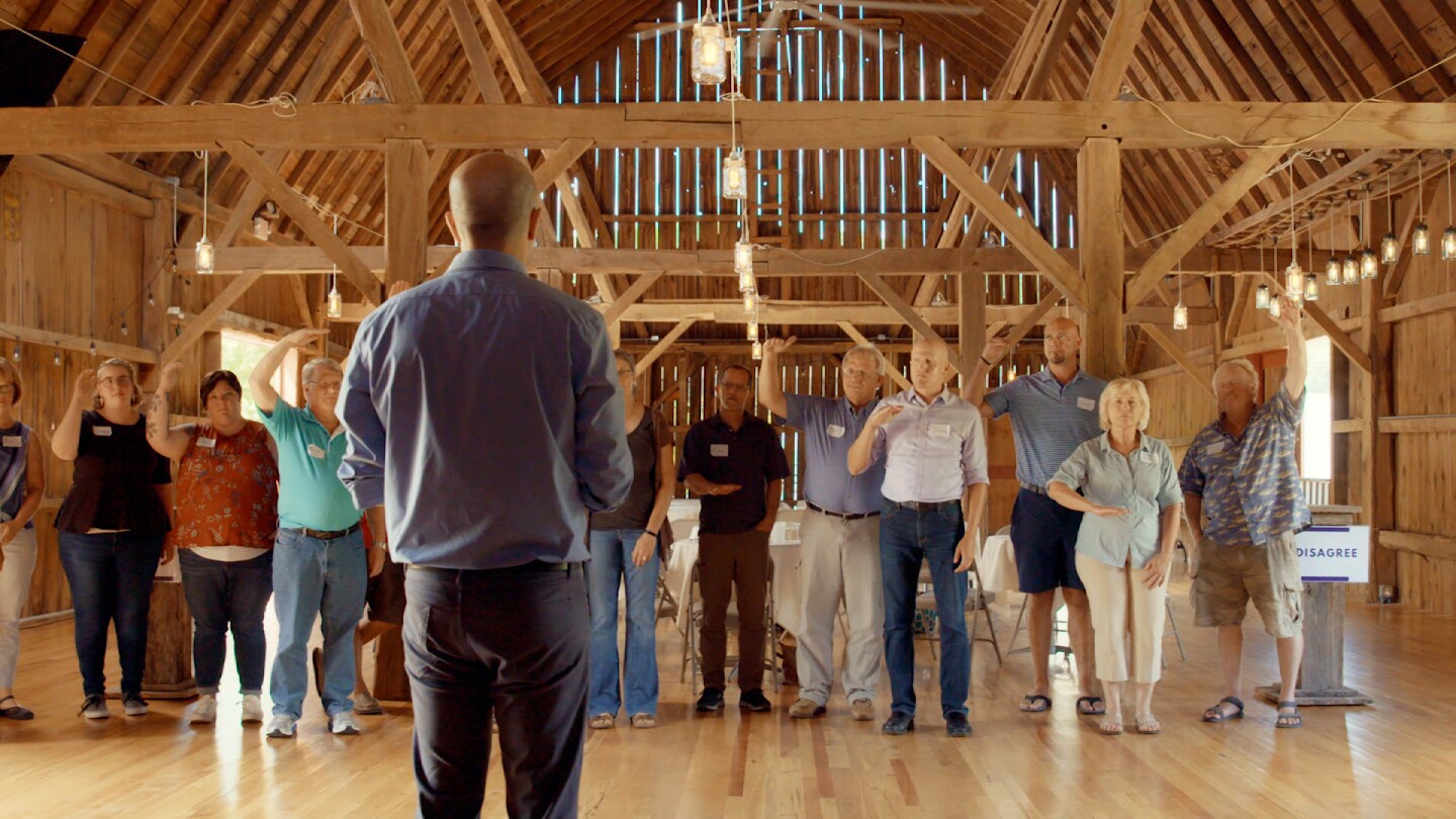 A group of people lined up are doing arm gestures in front of a man. | From "Purple"