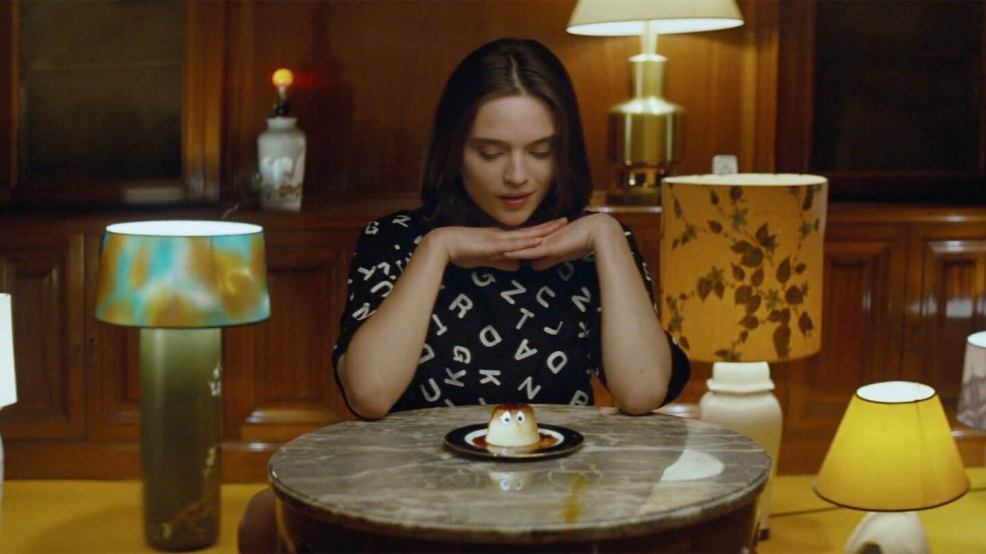 Still from Creme Caramel: A woman in a room with many lamps looks down at a creme caramel that has eyes.