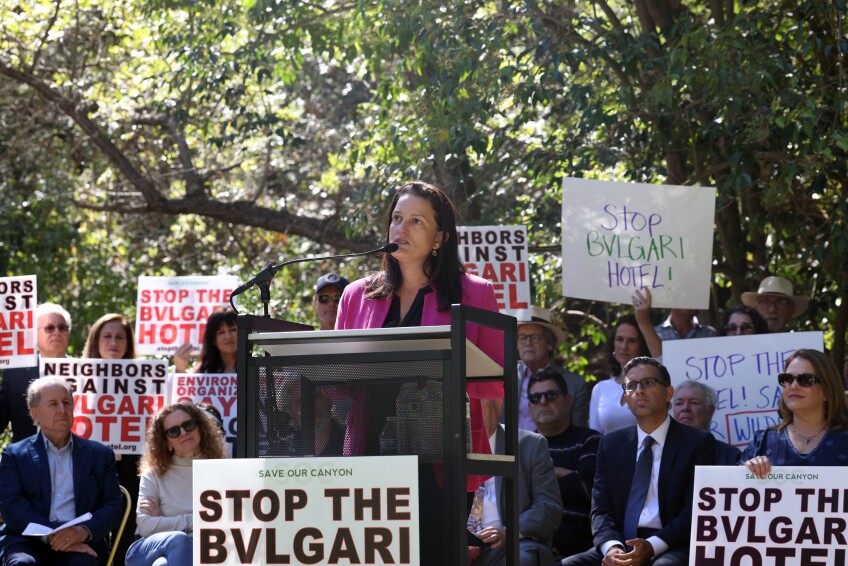 Katy Young Yaroslavsky speaks into a microphone behind a podium in front of a crowd holding signs that read "STOP THE BULGARI HOTEL" and "NEIGHBORS AGAINST BULGARI HOTEL"