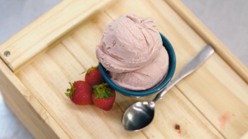 Strawberry ice cream in a small blue bowl displayed on a wooden tray with fresh strawberries and a spoon.