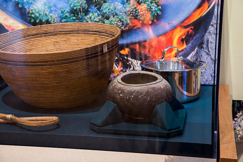 a Gabrielino/Tongva cooking basket and a Paiute cooking stick (just visible at the lower right), both from the early 20th century, and a Gabrielino/Tongva cooking bowl, sit beside a stainless steel cooking pot. Photo: The Autry Museum