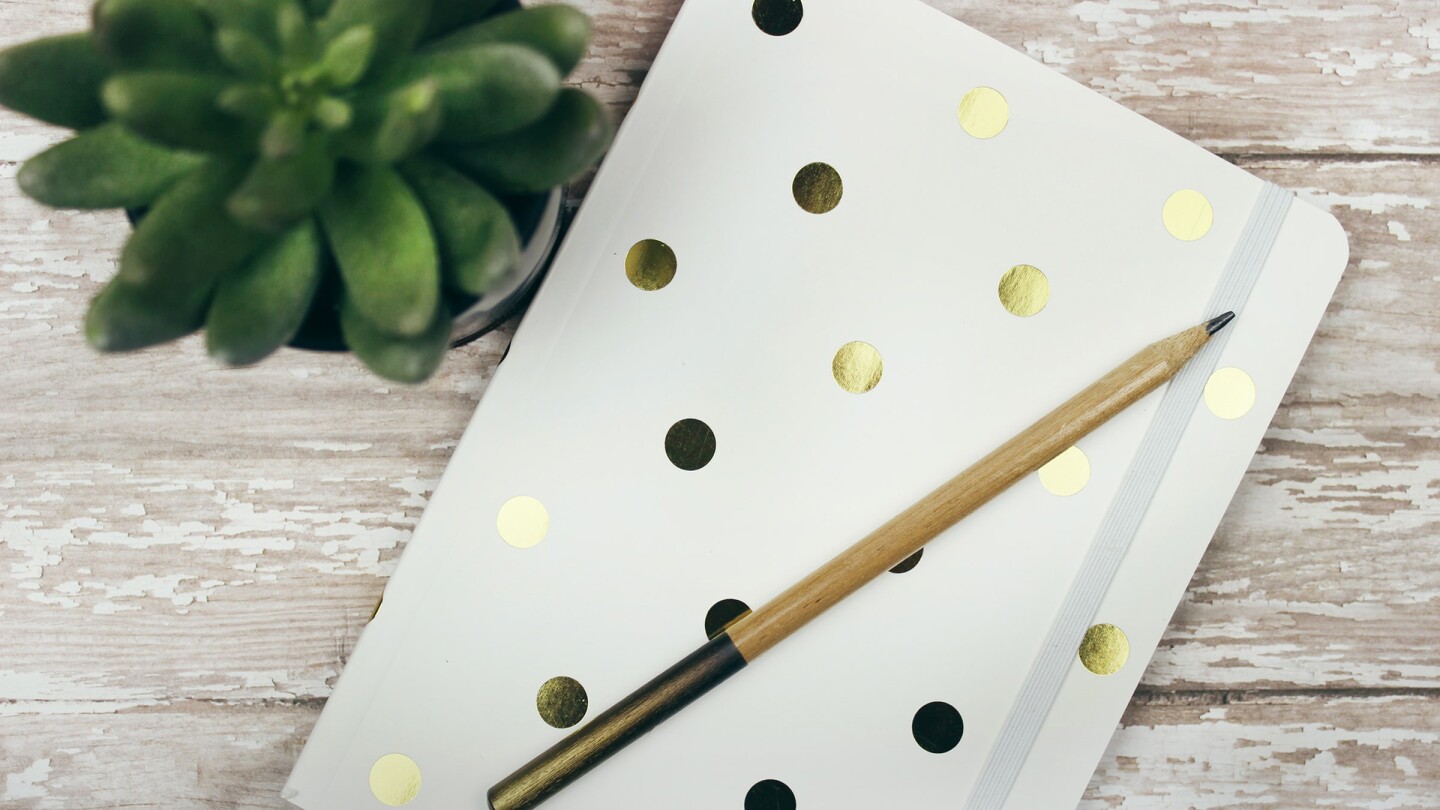 Gold and white polka dot notebook or journal sits on a white wooden desk with pencil and small planted succulent. | Jen Theodore / Unsplash