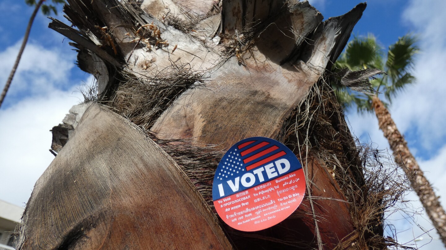 The trunk of a palm tree wears an "I Voted" sticker as it stands tall against a blue sky with white puffy clouds.
