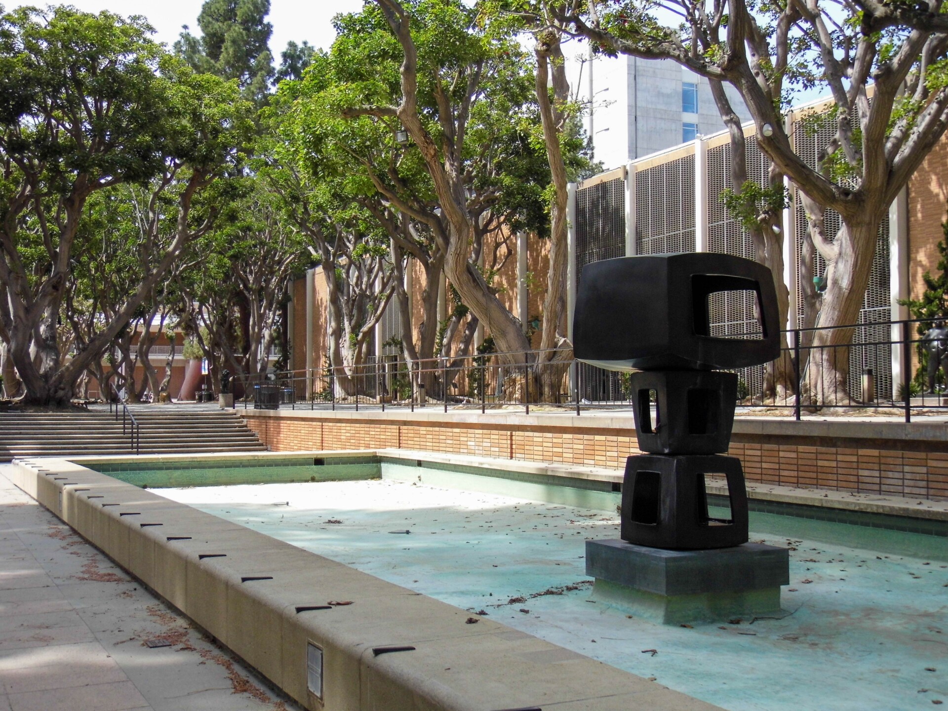 A black sculpture of three cube-geometric shapes stacked on top of one another, with the top cube being the largest. The cubes are asymmetrical and sit in an empty fountain amongst tall trees and campus buildings.