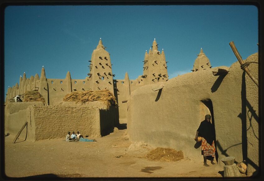 Tan colored mud mosque with three minarets topped by ostrich eggs with low buildings and people in foreground