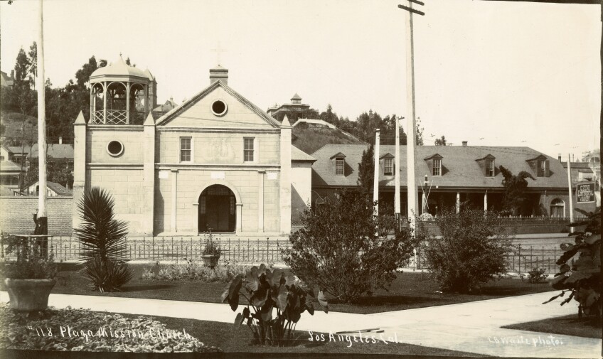 A black and white photo of a small church with a large arched doorway, two windows above it, and a pointed, A-frame roof. Next to the church is a bell tower, its bell encased with a decorative dome and archways.