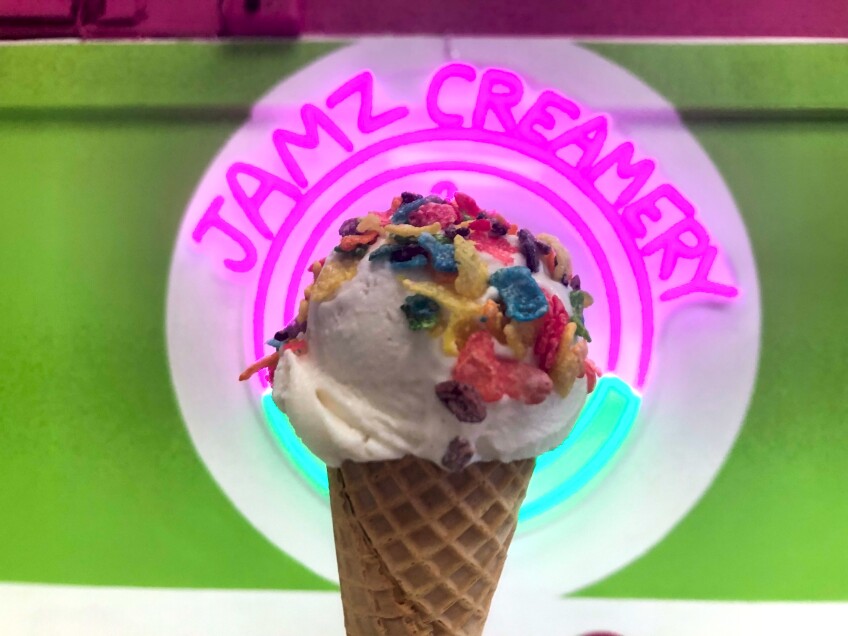 A scoop of white/cream-colored ice cream sits on top of a waffle cone. The ice cream is topped with multicolored popped rice cereal. Behind the ice cream is a lime green wall with a pink neon sign that reads, "Jamz Creamery." 